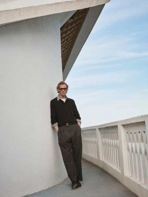 Adult male in a black sweater and grey slacks leans against a white wall and balustrade with a blue sky in the background.