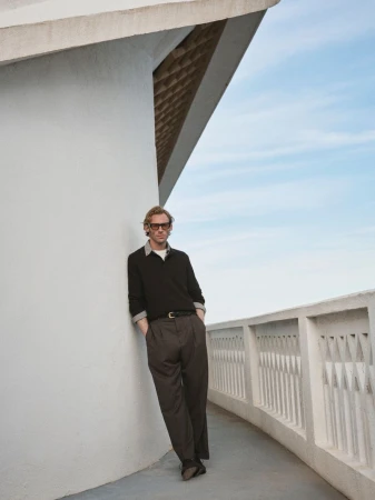 Adult male in a black sweater and grey slacks leans against a white wall and balustrade with a blue sky in the background.