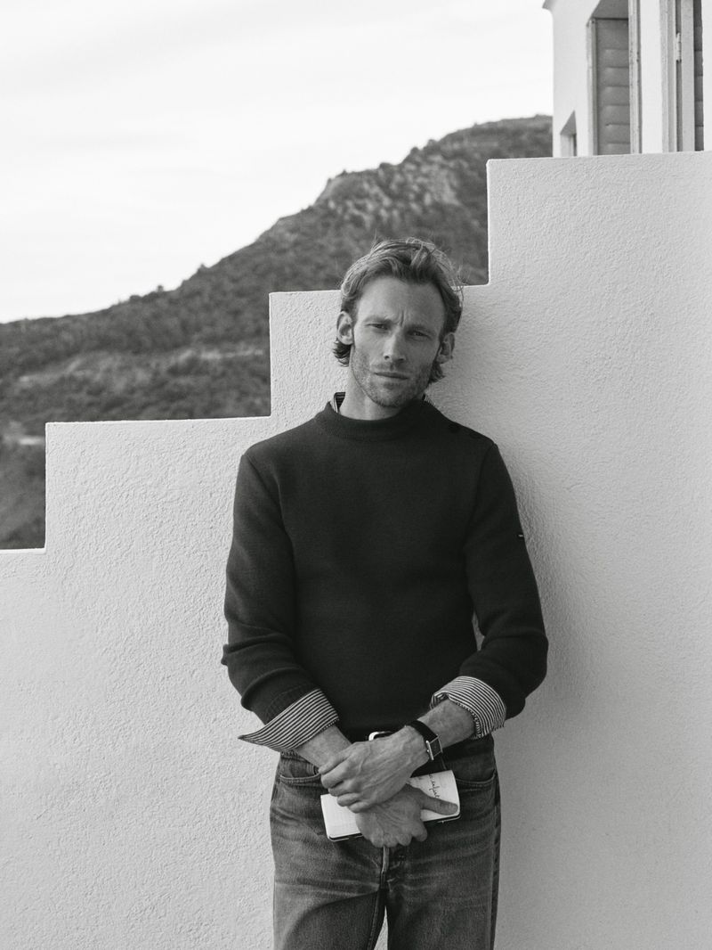 Black and white portrait of a man with a dark sweater and jeans, posing in front of a white wall, mountain visible in background.