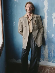 Full-length portrait of a male model leaning against a distressed blue wall wearing a beige blazer and dark trousers.