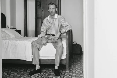 Black and white portrait of a man seated on a bed in a hotel room wearing a striped shirt and trousers.