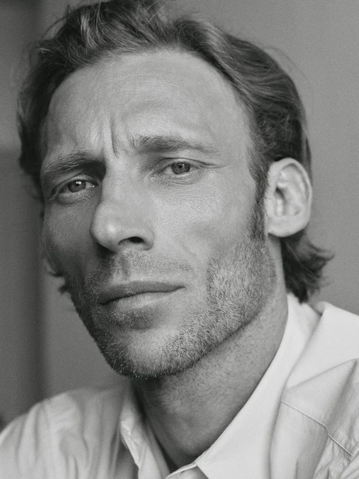 Close-up monochrome portrait of a man with stubble wearing a light-coloured collared shirt against a plain background