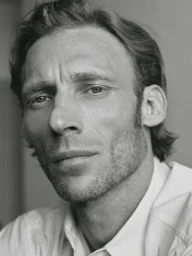 Close-up monochrome portrait of a man with stubble wearing a light-coloured collared shirt against a plain background