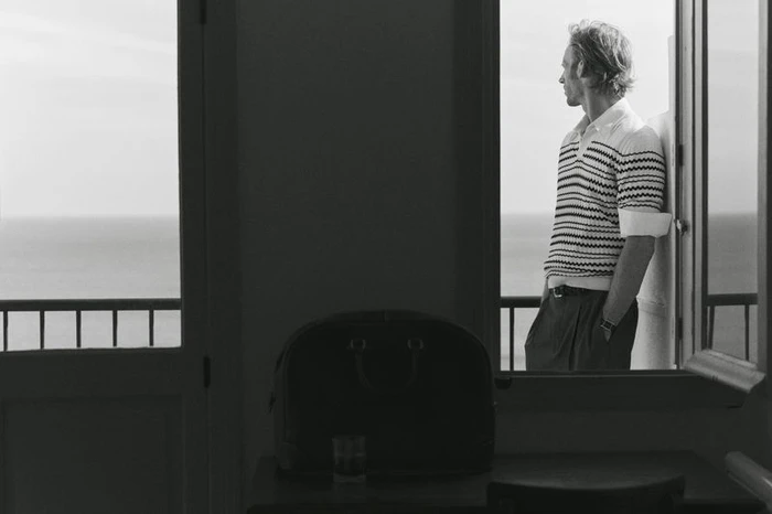 Man in striped polo shirt leaning on a balcony looking out toward the sea, photographed in black and white.