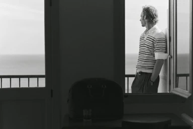 Man in striped polo shirt leaning on a balcony looking out toward the sea, photographed in black and white.