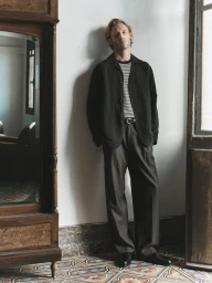 Man wearing a dark jacket over a striped t-shirt and trousers leaning against a white wall in an interior room.