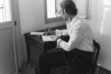 A man in a white shirt and dark trousers sitting at a wooden desk writing in a notebook in a bright room.
