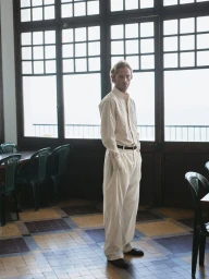 Man wearing cream-colored long-sleeve shirt and trousers standing inside a room with large grid windows and tiled floor.