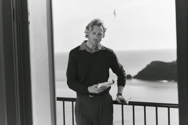 Black and white portrait of a man standing on a balcony holding a book with the sea in the background.