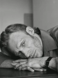 Portrait of a man resting his head on his hands, wearing a collared shirt and a dark wrist watch against a plain background