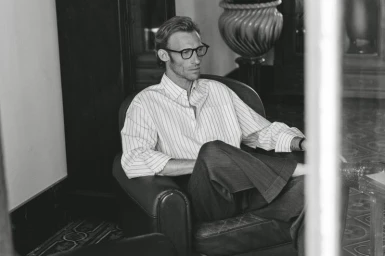 Man wearing eyeglasses and a striped button-down shirt seated in a leather armchair in a room.