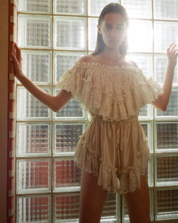 Elegant woman in a cream-colored, off-the-shoulder lace romper poses in front of a glass block wall, bathed in soft natural light.