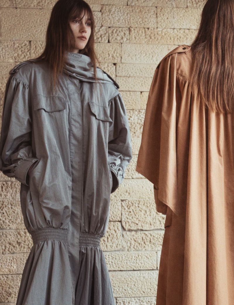 Two women wearing stylish grey and tan trench coats pose indoors against a textured brick wall in a fashion editorial shot.