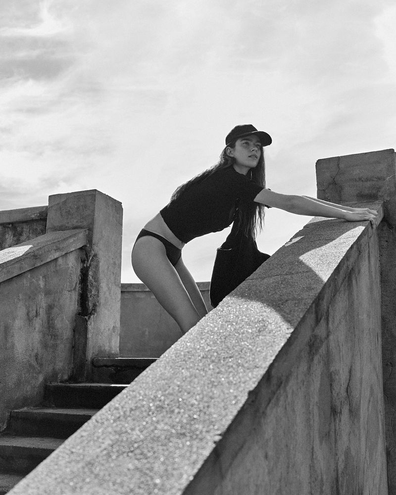 Young woman in a baseball cap poses on concrete stairs in a monochromatic outdoor setting, showcasing fashion and casual elegance.