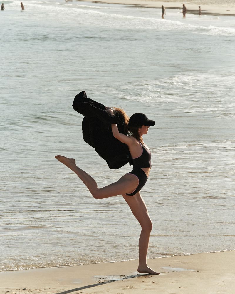 Woman in a black swimsuit and baseball cap leaps barefoot on a sandy beach with ocean waves and distant figures on a sunny day.