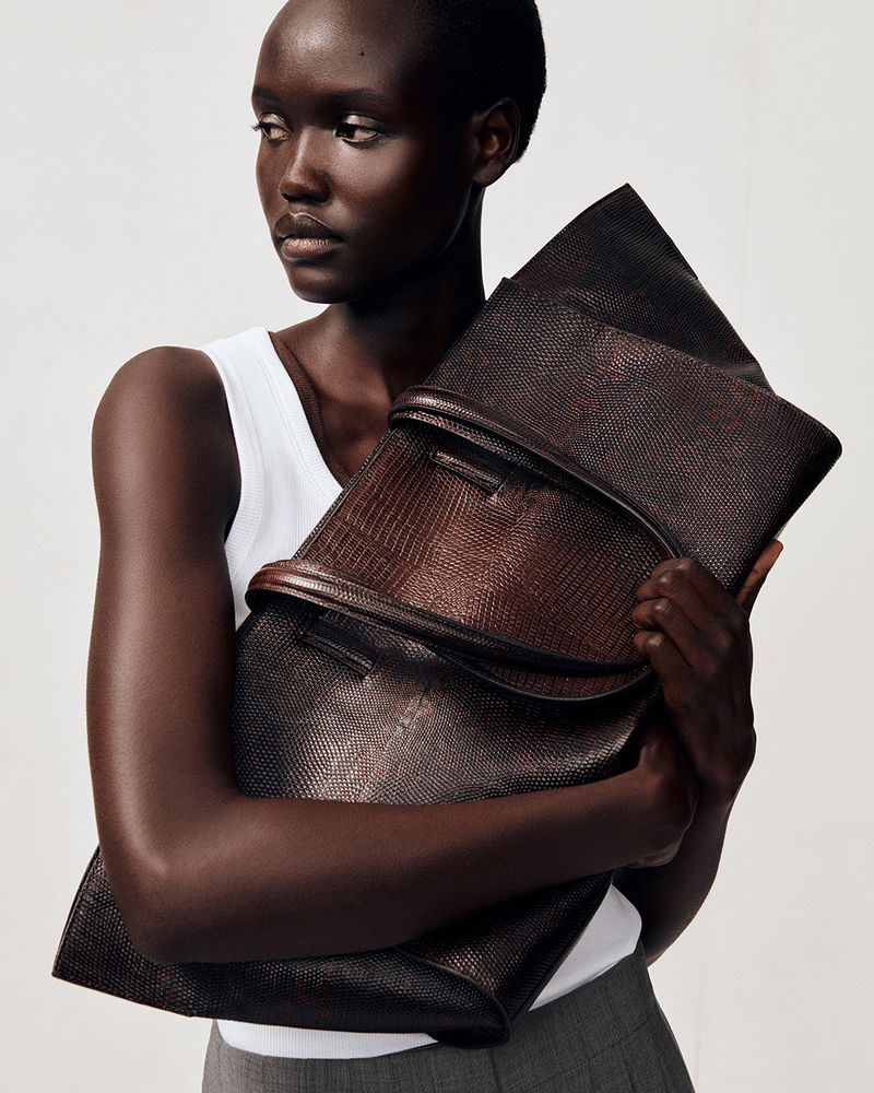Model wearing a white tank top and grey trousers holding a large dark brown textured leather clutch bag