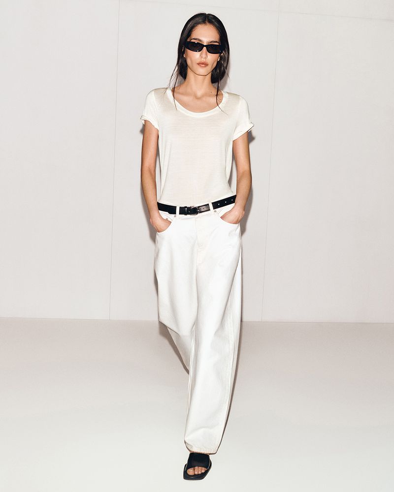A young adult woman with long dark hair walks toward the camera in a studio, wearing a white tee, baggy jeans, sandals, and sunglasses.