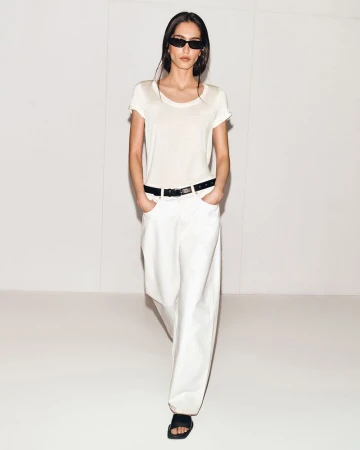 A young adult woman with long dark hair walks toward the camera in a studio, wearing a white tee, baggy jeans, sandals, and sunglasses.