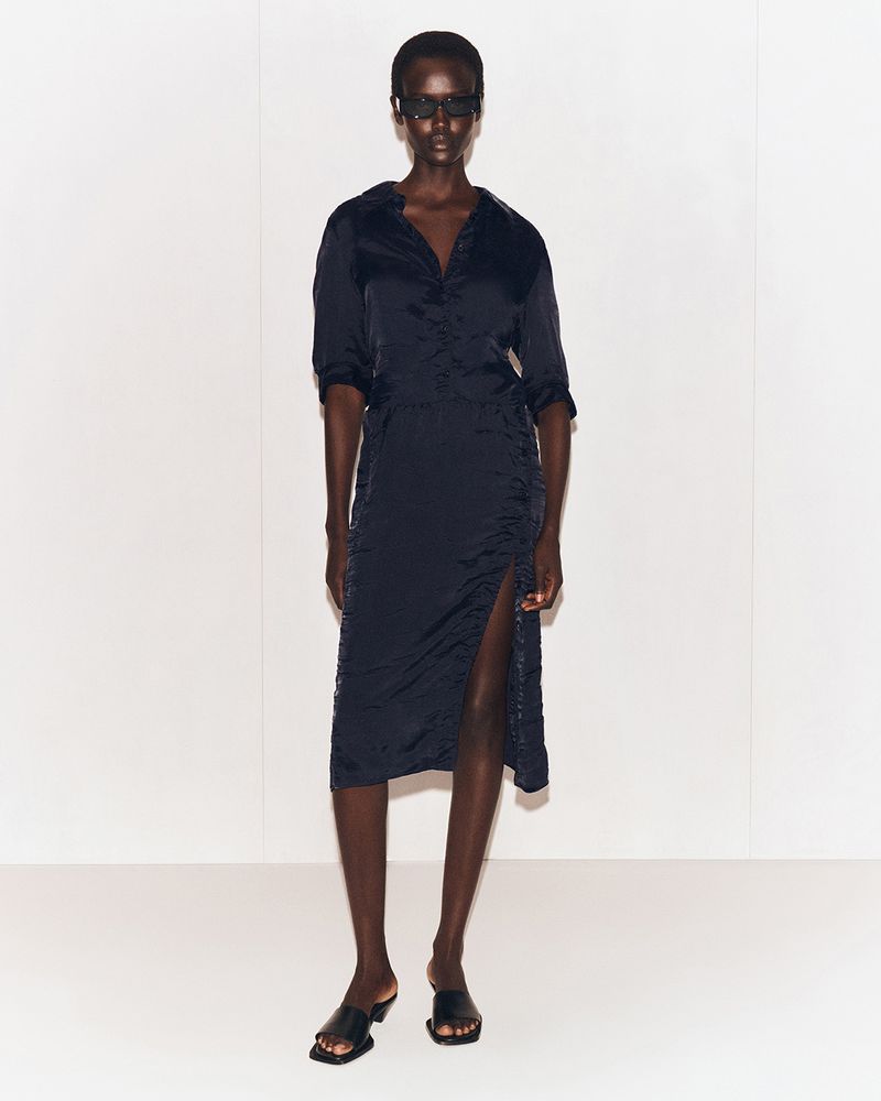 A black female adult poses in a navy satin midi dress with a side slit and black sandals against a bright white backdrop in a fashion studio.
