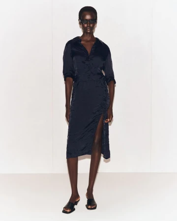A black female adult poses in a navy satin midi dress with a side slit and black sandals against a bright white backdrop in a fashion studio.