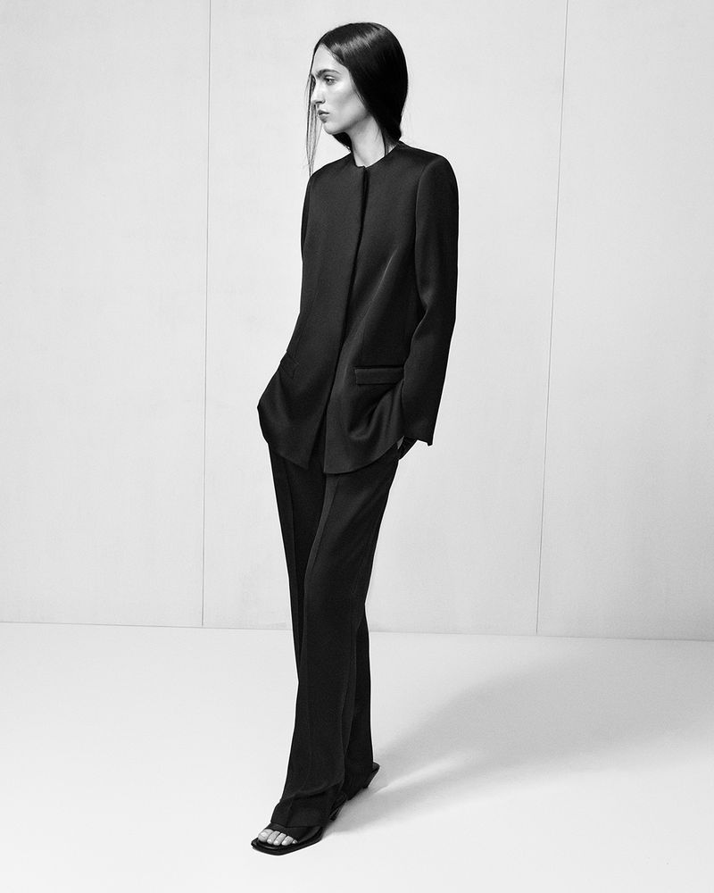 Monochrome fashion shot of a slender adult woman in a dark suit and minimalist sandals, posing against a white background.