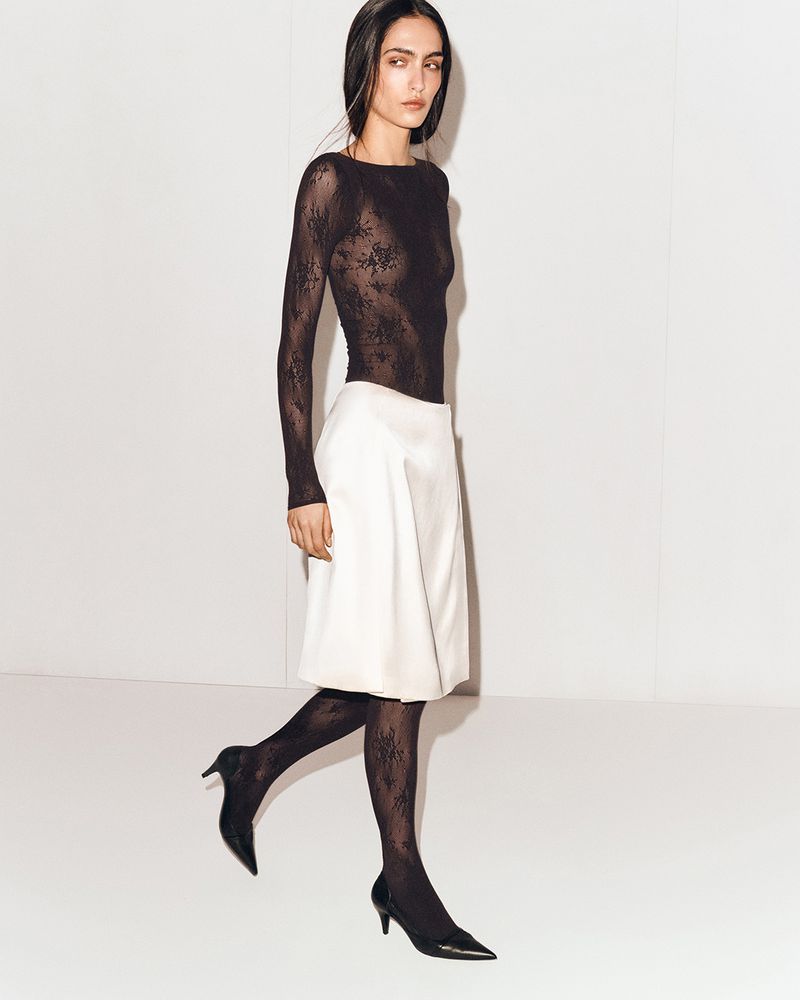 A young woman poses in a long-sleeved black lace top, white skirt, and black floral tights with kitten heels against a white backdrop.