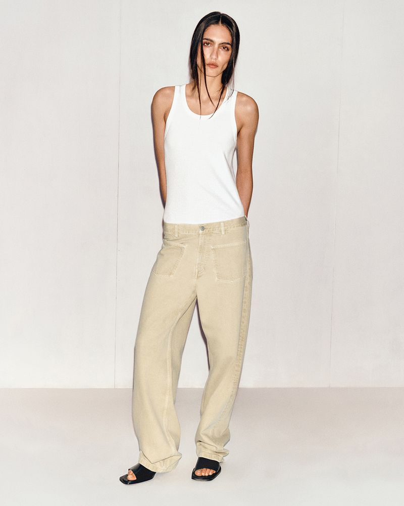 Adult woman in a white tank top and loose tan pants stands in a photo studio with a white background, looking at the camera.
