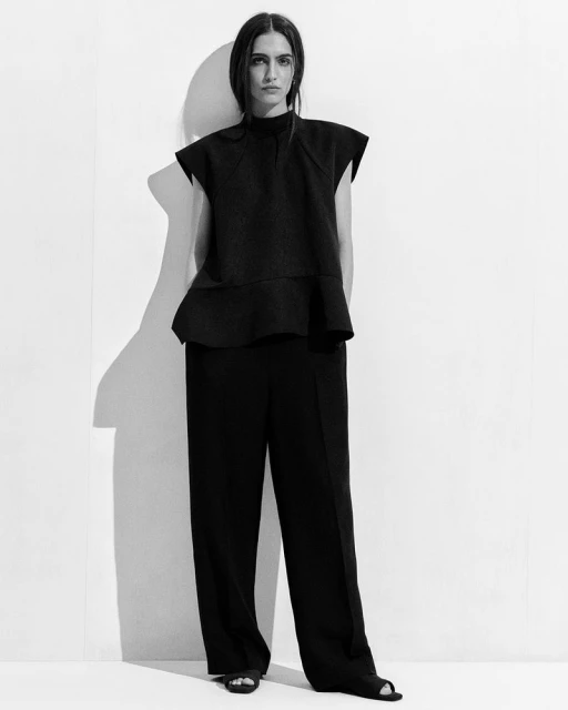 Full-length shot of a woman in a black top and wide-legged trousers standing against a white wall, casting a soft shadow.