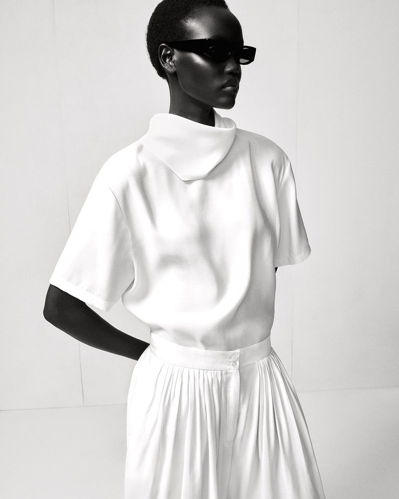 Monochromatic fashion shot featuring an adult female in a white short-sleeved top and pleated skirt, wearing dark sunglasses against a white backdrop.