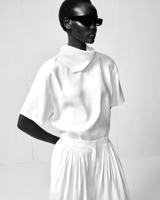 Monochromatic fashion shot featuring an adult female in a white short-sleeved top and pleated skirt, wearing dark sunglasses against a white backdrop.