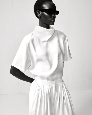 Monochromatic fashion shot featuring an adult female in a white short-sleeved top and pleated skirt, wearing dark sunglasses against a white backdrop.