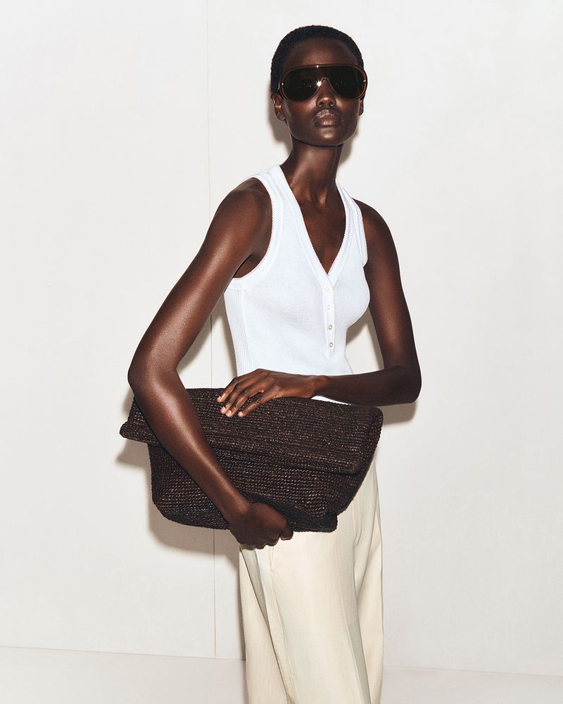 Dark-skinned adult female fashion model poses with large brown clutch bag in a minimalist photo studio setting. She wears sunglasses.
