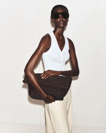 Dark-skinned adult female fashion model poses with large brown clutch bag in a minimalist photo studio setting. She wears sunglasses.