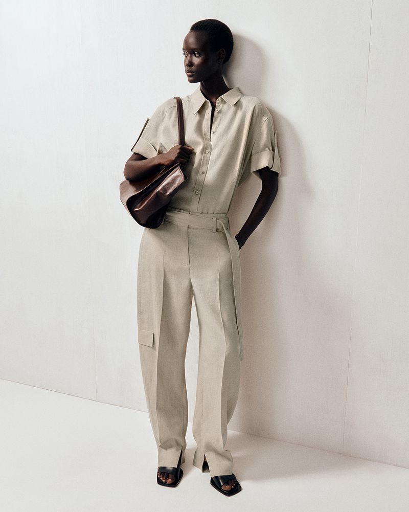 Editorial fashion shot of a Black female model wearing a light beige linen shirt and wide leg pants, carrying a brown leather shoulder bag.