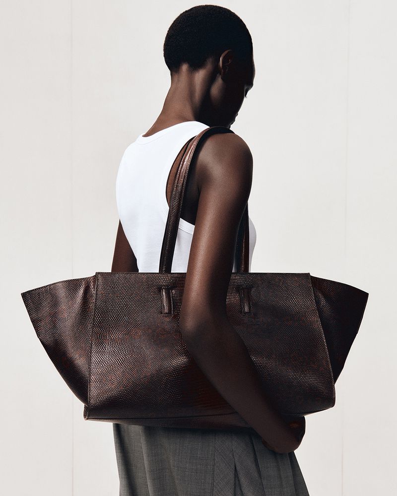 Dark-skinned female model carries a textured brown leather tote bag, wearing a white top and grey skirt against a white backdrop.