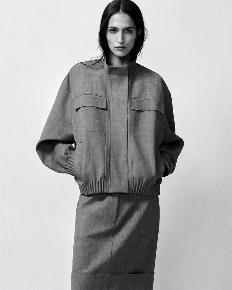 Black and white fashion shot of a woman posing indoors wearing a gray skirt suit with jacket and skirt, set against a white backdrop.