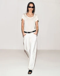 Model wearing dark sunglasses, a white t-shirt, and white trousers standing against a white seamless background.