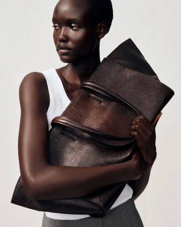 Model wearing a white tank top and grey trousers holding a large dark brown textured leather clutch bag