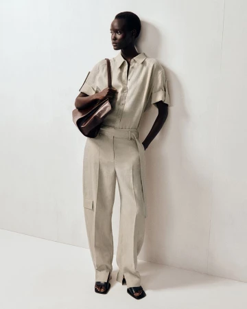 Model in beige short-sleeve shirt and matching trousers holding a dark leather shoulder bag against a white wall.