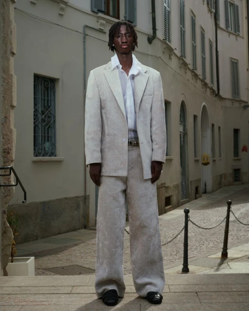 Man poses in an elegant off-white suit against an old European city street backdrop. Fashion editorial showcasing modern tailoring.