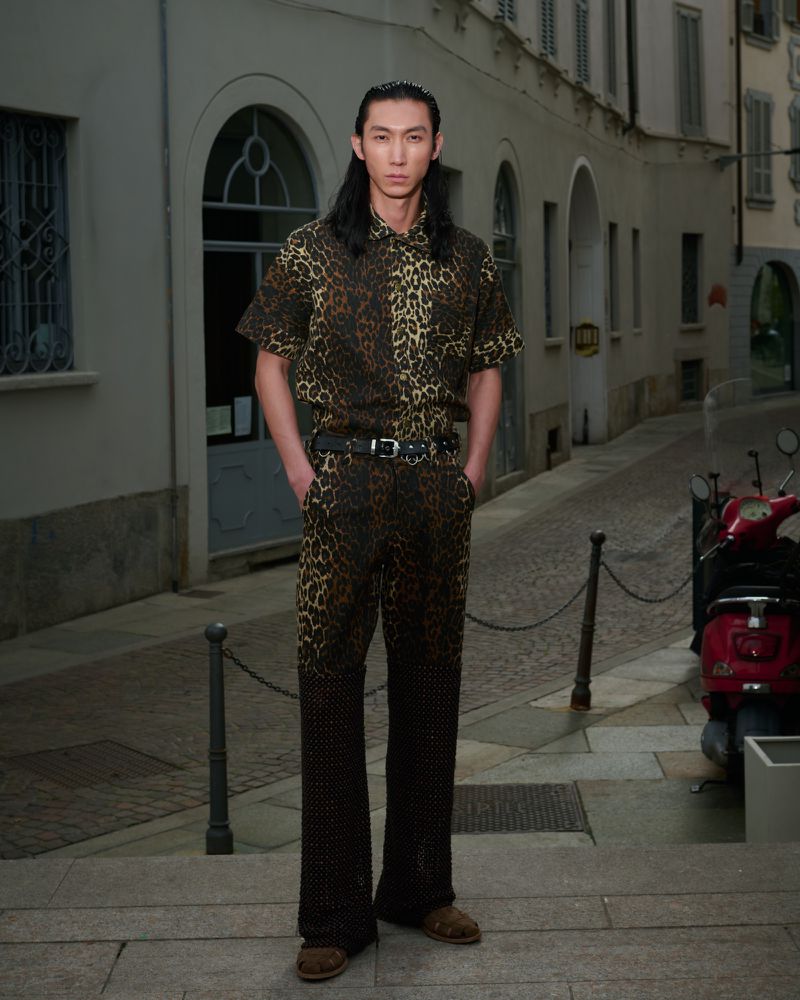 Man poses in leopard-print shirt and pants on city street with scooter. He has long dark hair and wears a black belt and brown shoes.