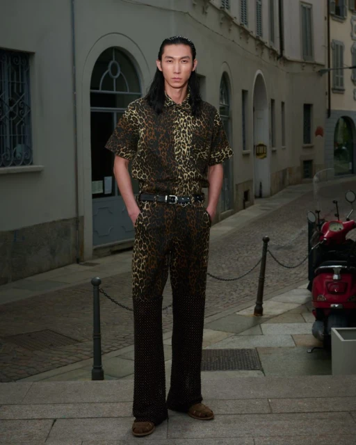 Man poses in leopard-print shirt and pants on city street with scooter. He has long dark hair and wears a black belt and brown shoes.