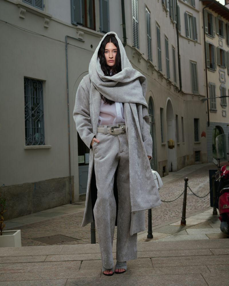 A solitary figure stands on weathered steps in an urban setting, draped in a long grey hooded coat, paired with matching trousers.