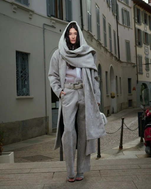 A solitary figure stands on weathered steps in an urban setting, draped in a long grey hooded coat, paired with matching trousers.