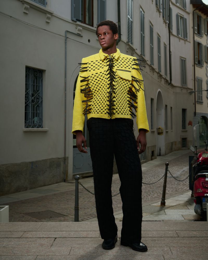 Adult male poses in a yellow and black jacket with black pants in an urban environment with buildings and a scooter visible.