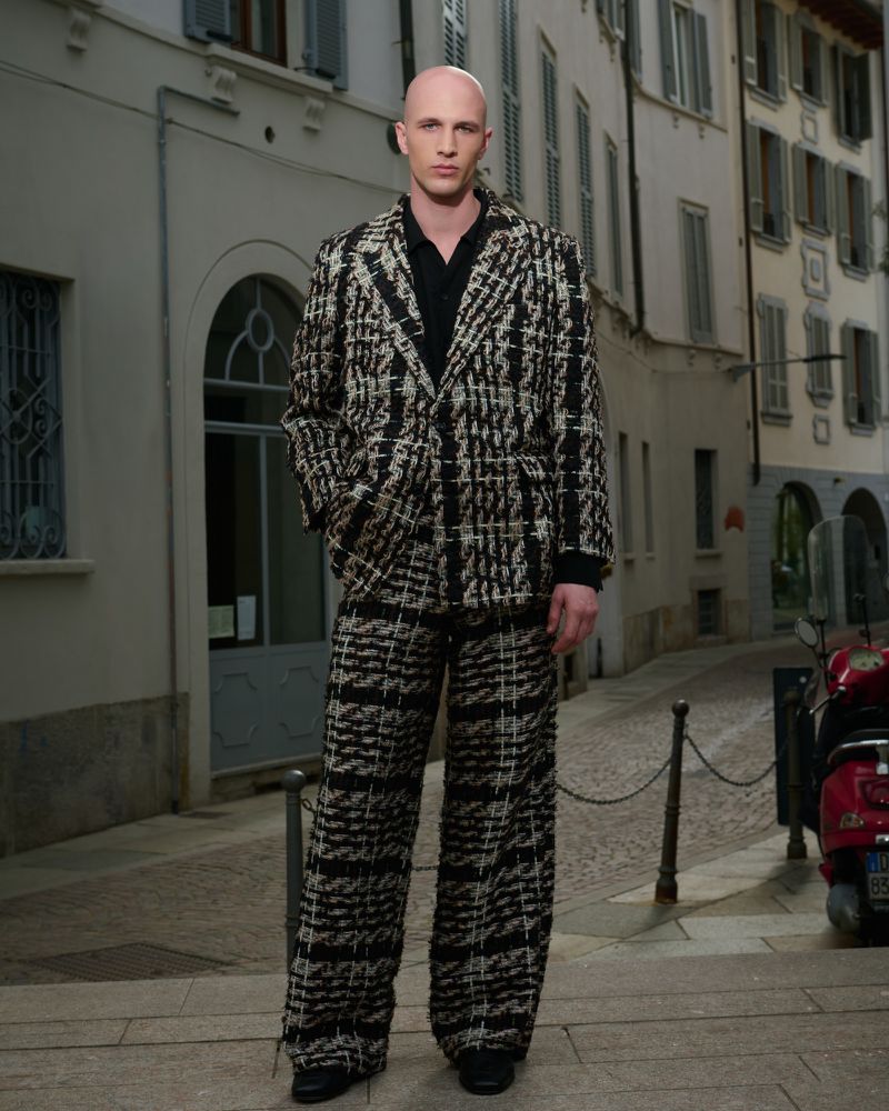A bald man in a patterned tweed suit stands on a cobblestone street with buildings and a scooter visible in the background.