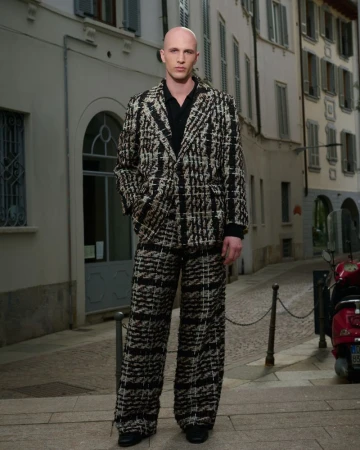 A bald man in a patterned tweed suit stands on a cobblestone street with buildings and a scooter visible in the background.