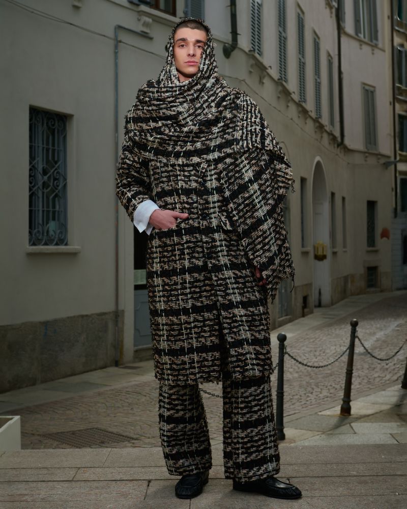 Full-length portrait of a fashion model in tweed-like coat and pants with matching scarf draped over his head in an urban outdoor setting.