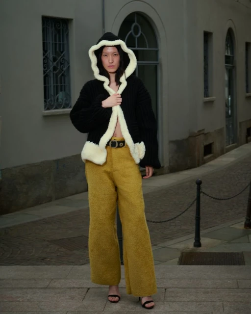 Fashionable woman poses in a black hooded jacket with cream trim and mustard corduroy pants on an urban sidewalk with neutral walls.
