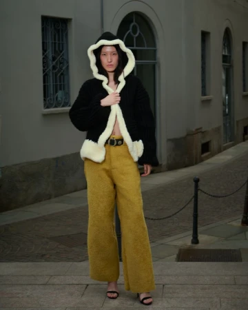 Fashionable woman poses in a black hooded jacket with cream trim and mustard corduroy pants on an urban sidewalk with neutral walls.